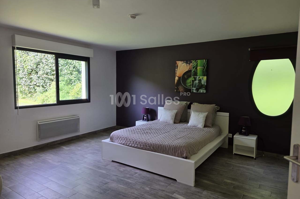 Chambre moderne avec lit double, mur violet, tableau décoratif, grande fenêtre et sol en carrelage gris.