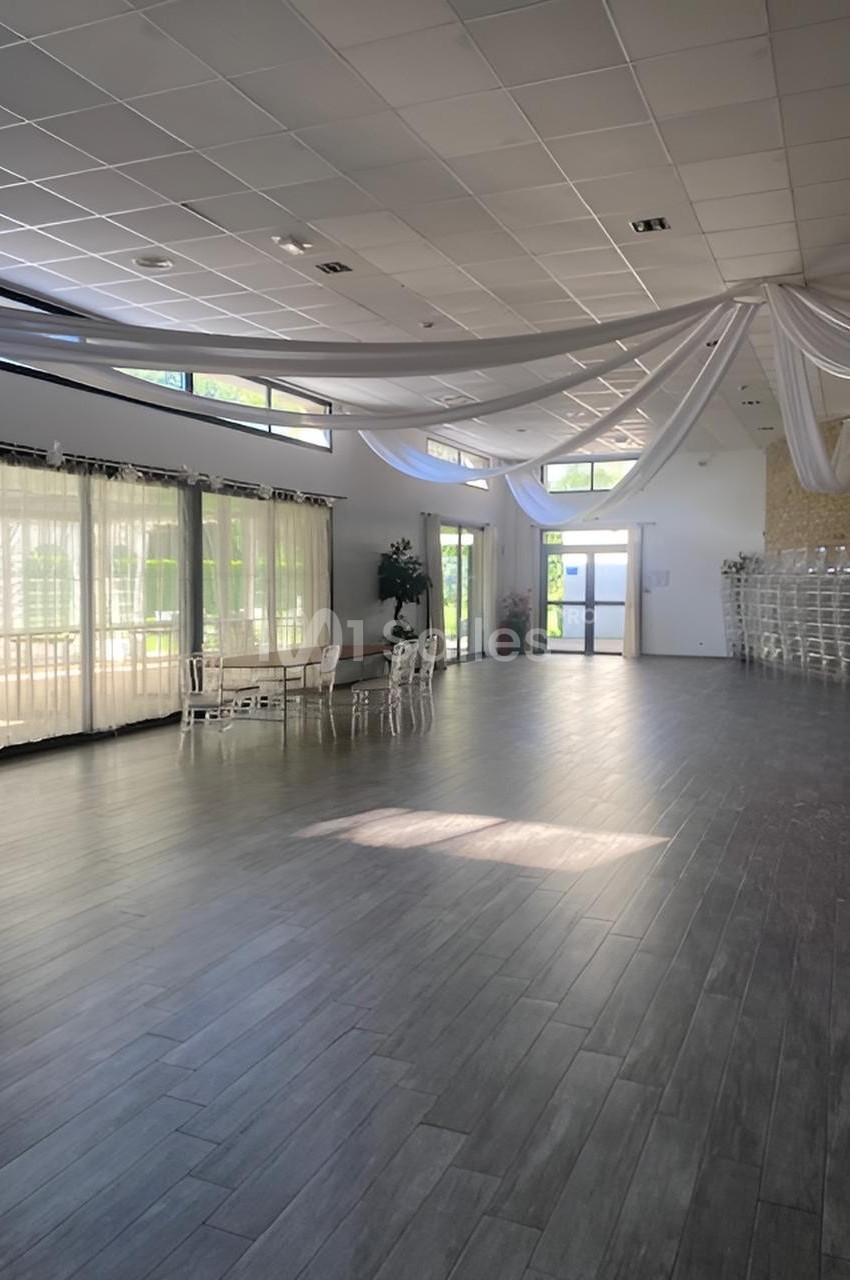 Salle spacieuse avec parquet sombre, grandes baies vitrées, plafond décoré de voilages blancs et chaises empilées.