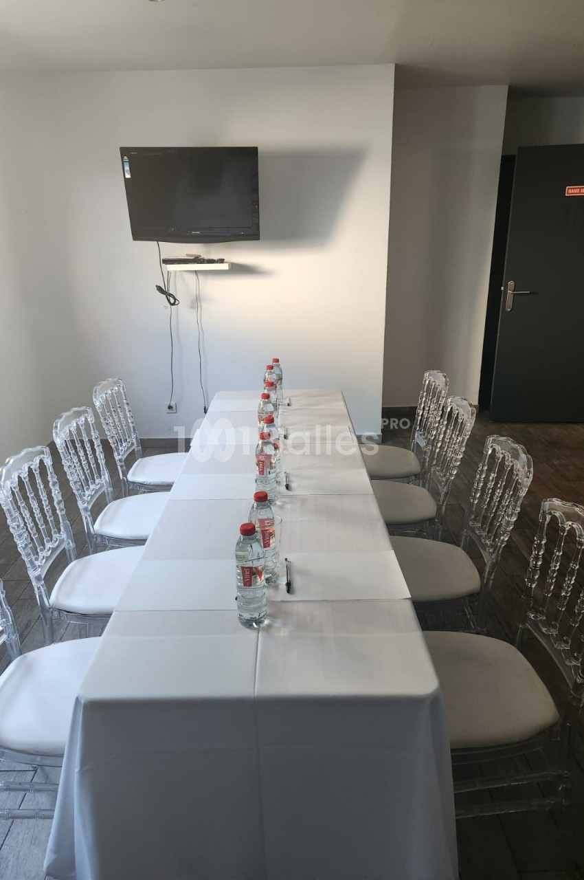 Salle de réunion avec une table rectangulaire couverte d'une nappe blanche, chaises transparentes et écran mural.