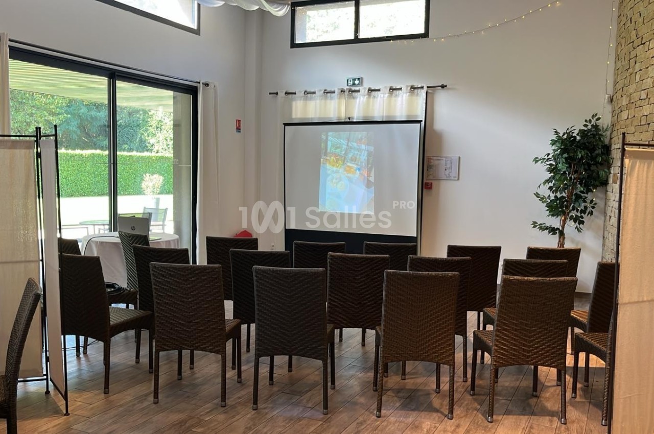 Salle aménagée avec des chaises en rotin face à un écran de projection, éclairée par la lumière naturelle d'une baie vitrée.