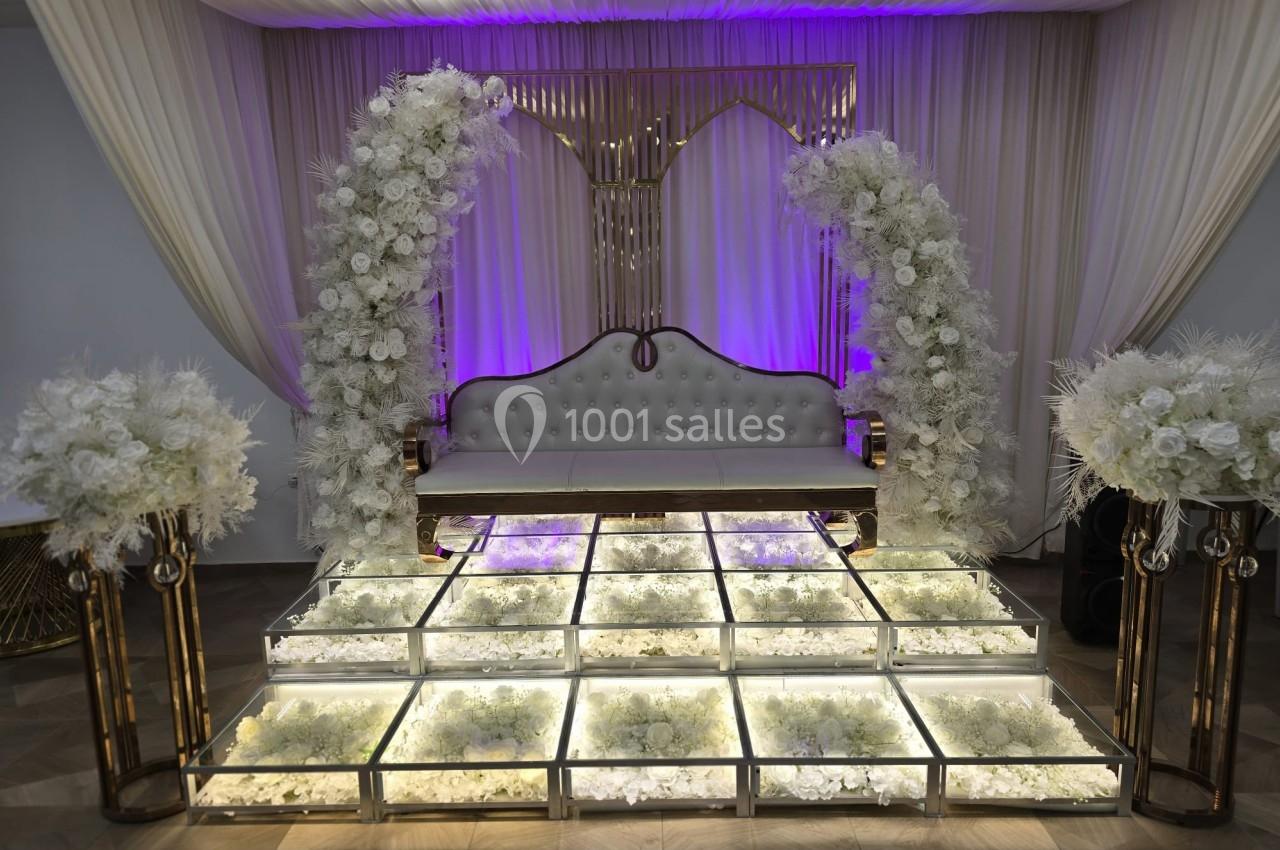 Canapé blanc entouré de décorations florales blanches, posé sur une estrade lumineuse dans une salle ornée de rideaux.