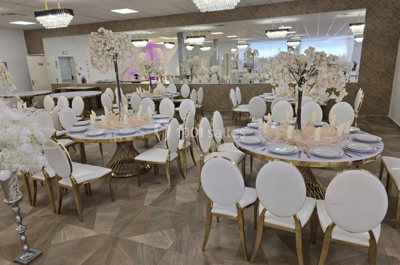 Salle de réception élégante avec tables rondes décorées, chaises blanches et lustres suspendus.