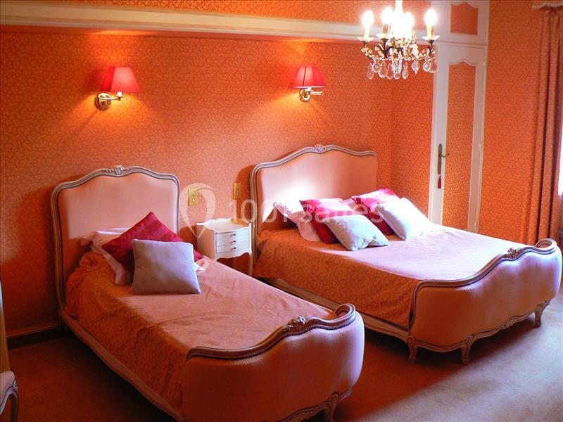 Deux lits simples avec têtes de lit roses dans une chambre aux murs orange, éclairée par un lustre et des appliques murales.