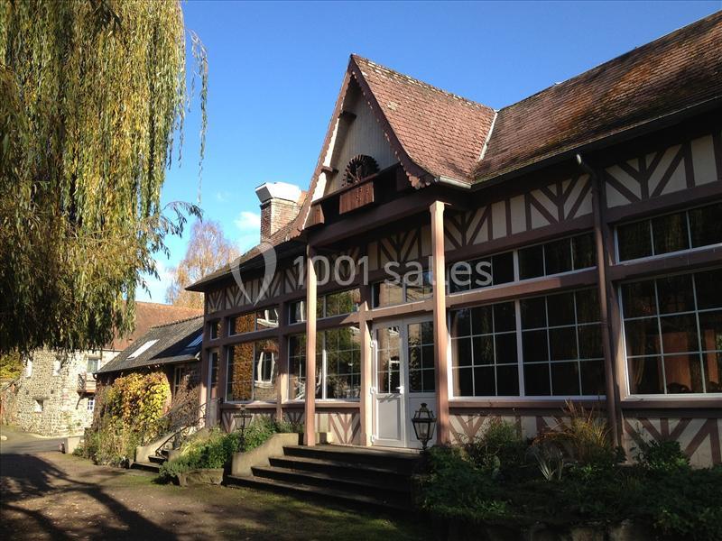 Façade d'une maison à colombages avec de grandes fenêtres, entourée de végétation sous un ciel bleu.