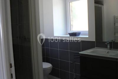 Salle de bain moderne avec carrelage gris, lavabo, toilettes et fenêtre laissant entrer la lumière naturelle.