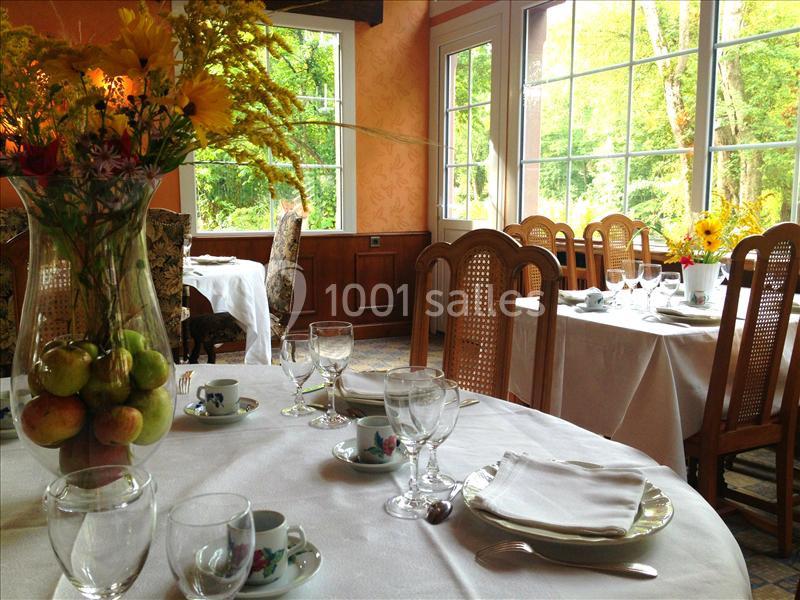 Salle de restaurant lumineuse avec tables dressées, nappes blanches, vaisselle élégante et vue sur un jardin verdoyant.