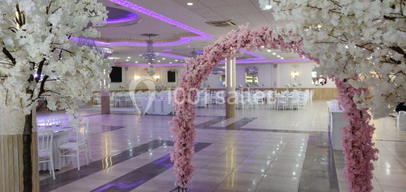 Salle de réception décorée avec des arches fleuries roses et blanches, tables dressées et éclairage violet.