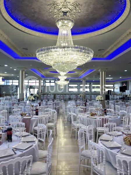 Salle de réception élégante avec tables dressées, chaises blanches et grands lustres en cristal illuminés.