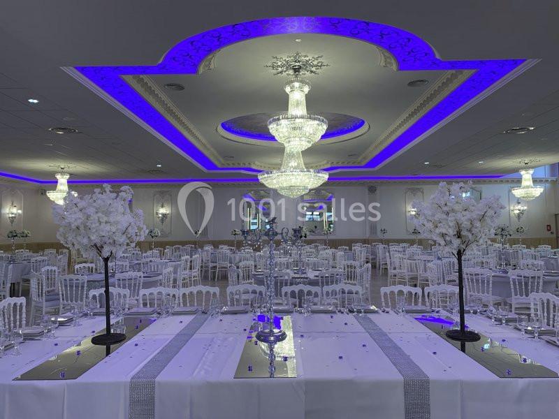 Salle de réception élégante avec tables dressées, chandeliers, décorations florales blanches et éclairage violet.