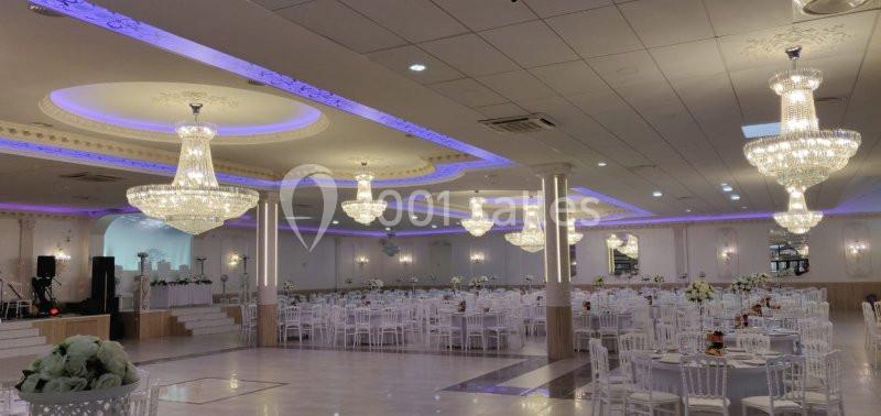 Salle de réception spacieuse avec tables rondes, chaises blanches et lustres en cristal suspendus au plafond.