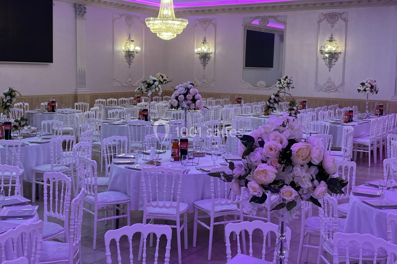 Salle de réception élégante avec tables rondes dressées, décorations florales et éclairage violet au plafond.