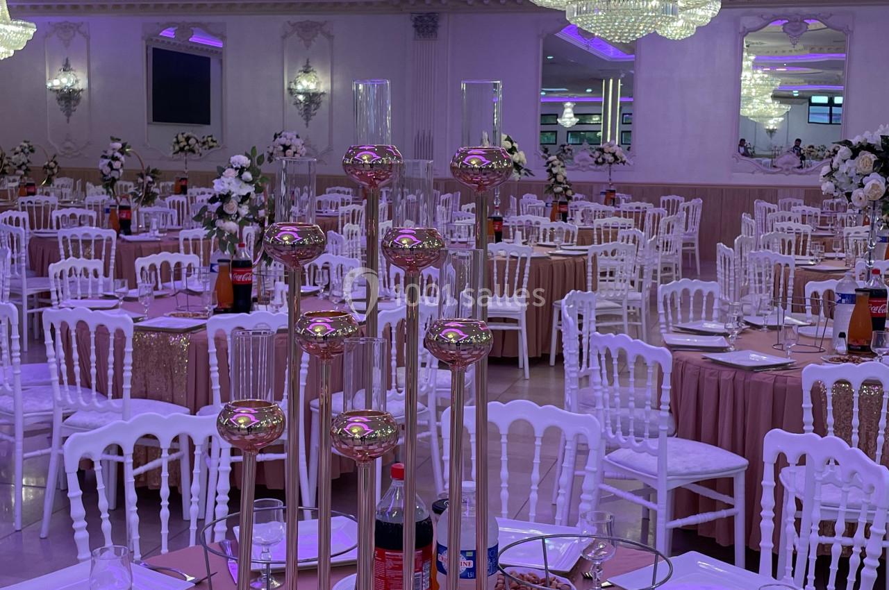 Salle de réception élégante avec tables dressées, nappes roses, chandeliers dorés et lustres en cristal lumineux.
