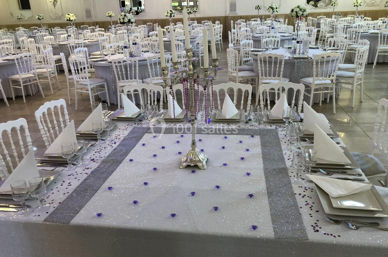 Salle de réception décorée avec des tables dressées, nappes scintillantes, chandeliers et chaises blanches.