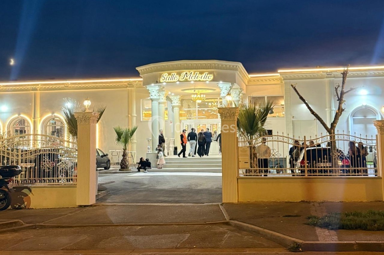 Façade illuminée d'une salle de réception en soirée, avec des invités rassemblés près de l'entrée.