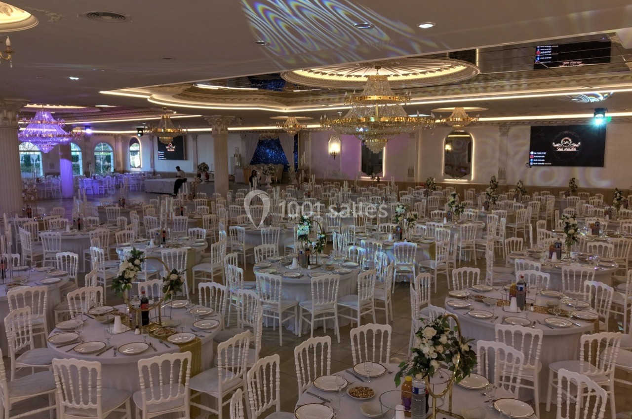 Salle de réception élégante avec de nombreuses tables rondes dressées, chaises blanches et lustres suspendus.