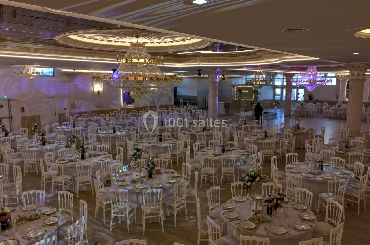 Salle de réception élégante avec tables rondes dressées, chaises blanches et lustres imposants au plafond.