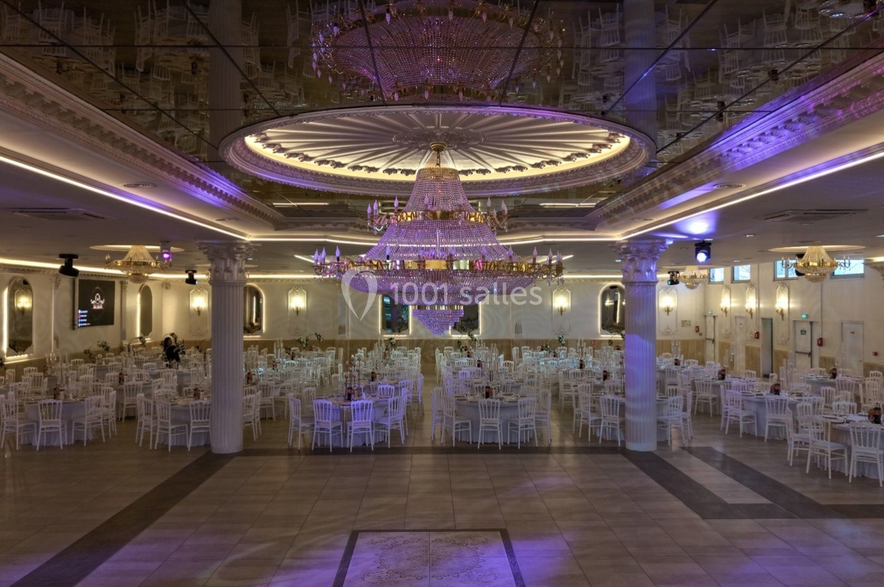 Salle de réception élégante avec un grand lustre central, des tables rondes et des chaises blanches disposées symétriquement.