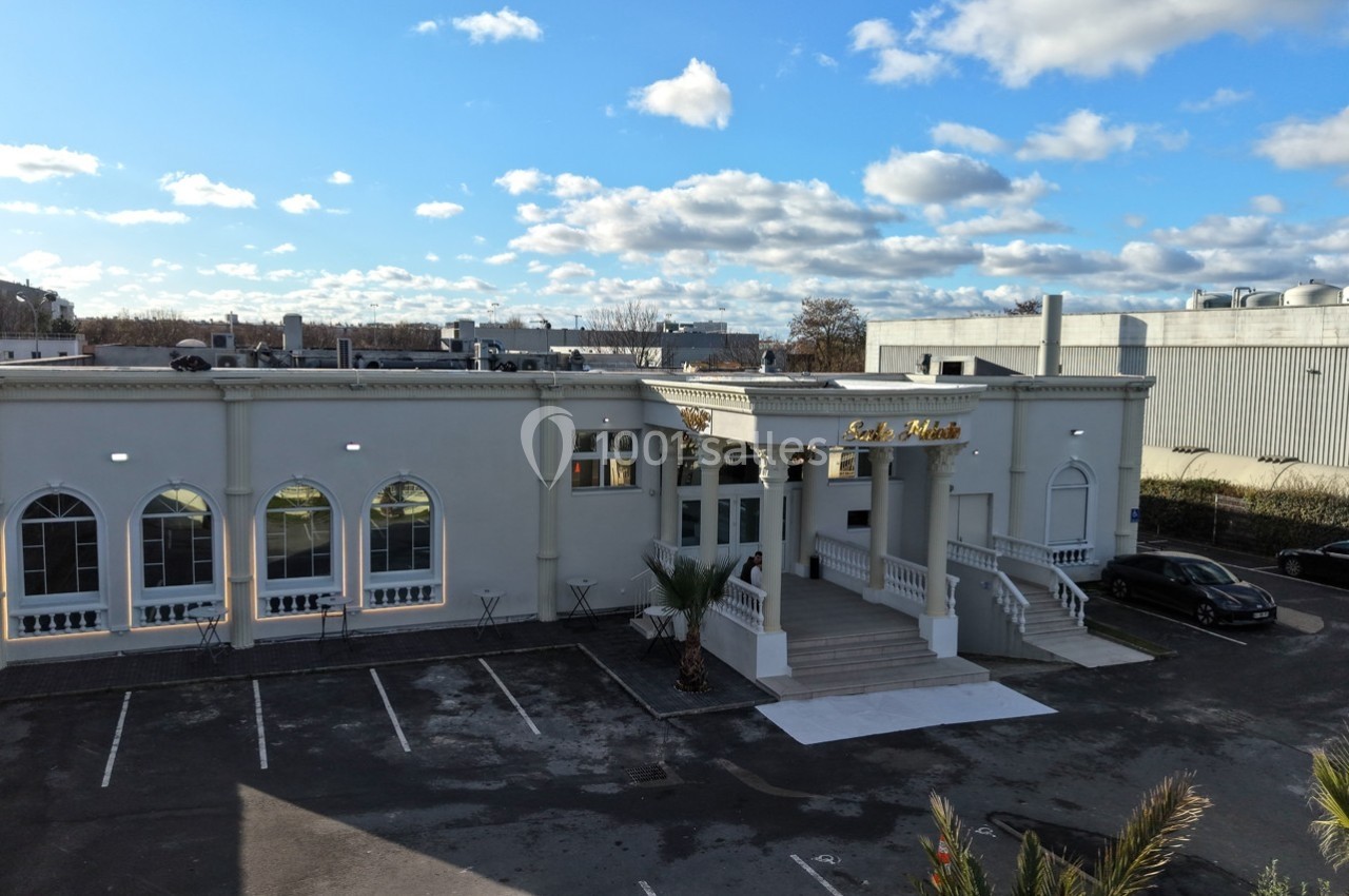 Façade d'un bâtiment blanc avec des arches vitrées, un escalier central et un parking vide à l'avant.