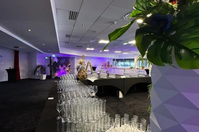 Salle de réception avec tables dressées, verres alignés au premier plan et décorations florales en arrière-plan.