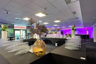 Salle de réception avec tables dressées, verres alignés au premier plan et décorations florales en arrière-plan.