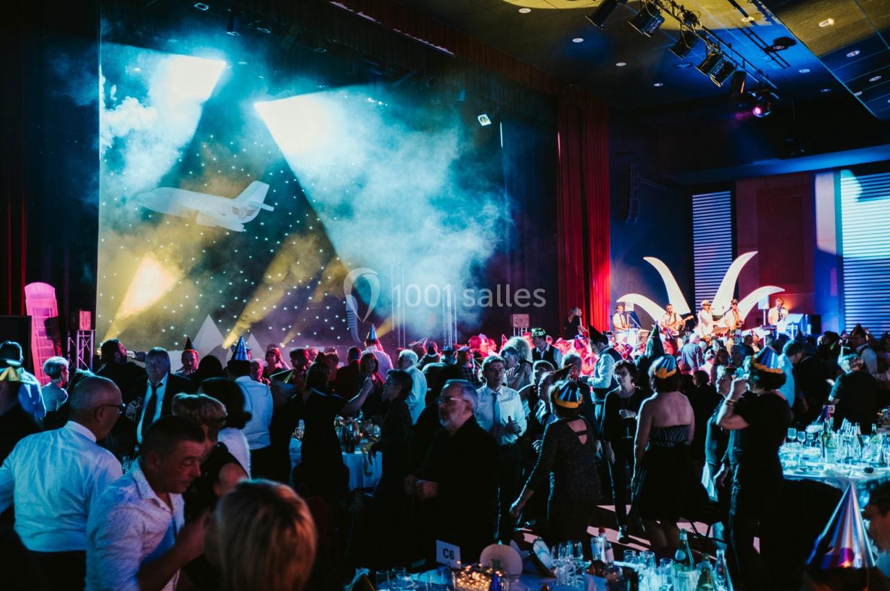 Salle de réception animée avec des invités debout, éclairages colorés et une scène en arrière-plan.