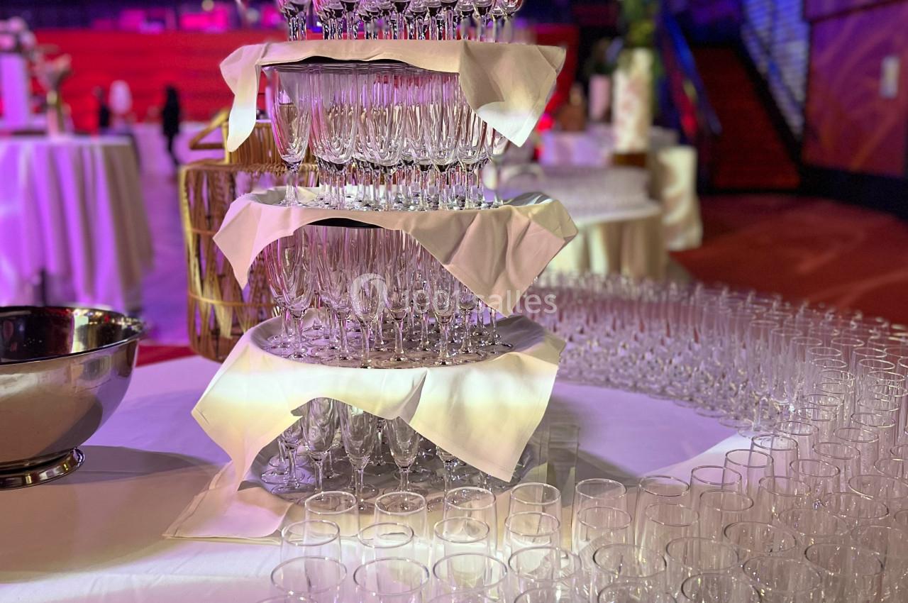 Tour de flûtes à champagne empilées avec des serviettes blanches, entourée de tables et de verres supplémentaires.