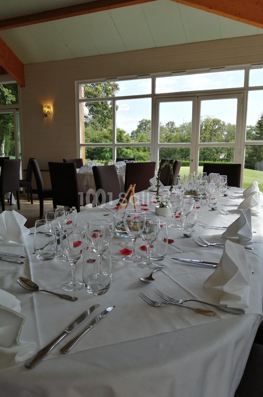 Table élégamment dressée avec nappes blanches, verres, couverts et décorations florales, dans une salle lumineuse avec vue…
