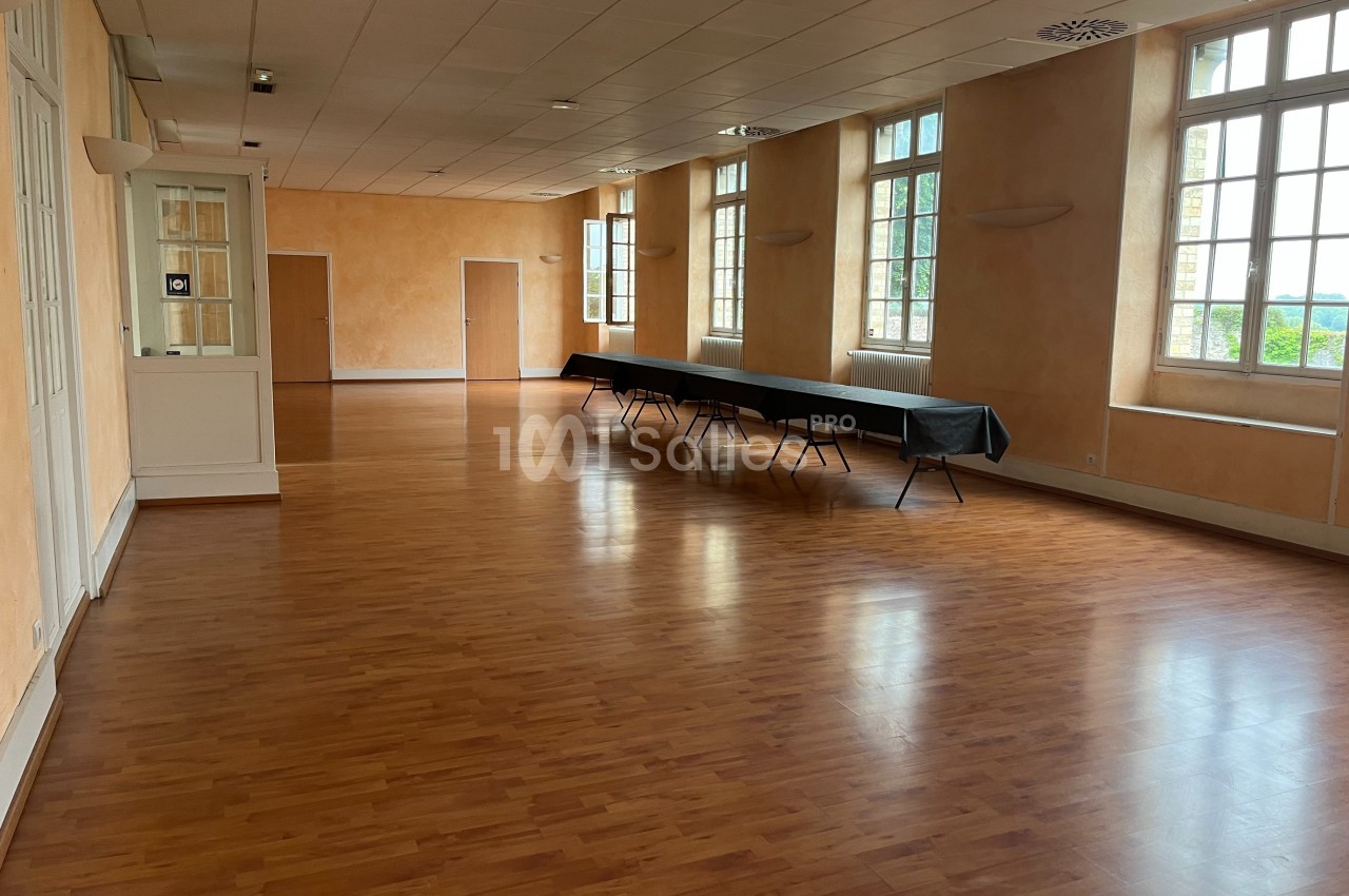 Salle vide avec parquet, grandes fenêtres laissant entrer la lumière naturelle et une table pliante noire le long du mur.