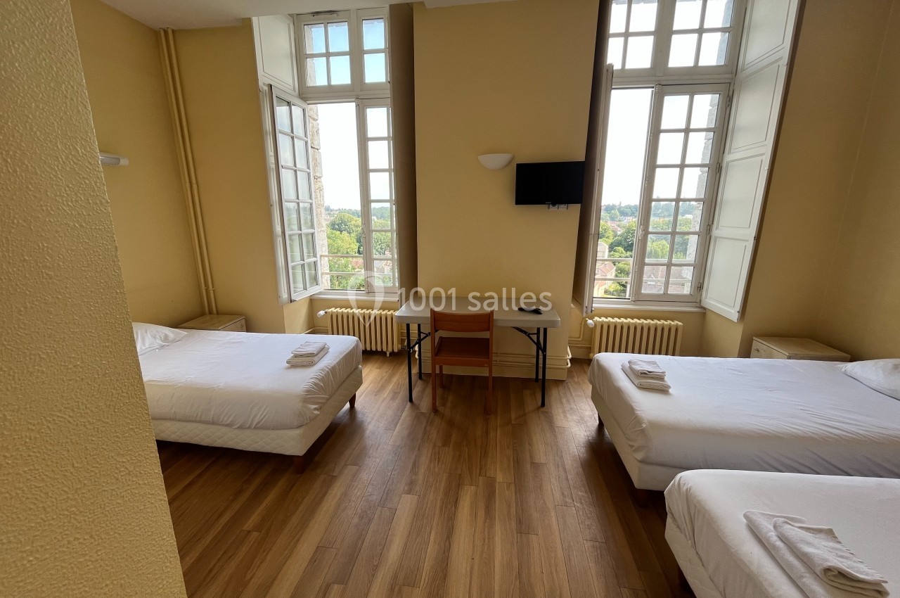 Chambre lumineuse avec trois lits simples, un bureau, une télévision murale et de grandes fenêtres donnant sur un paysage…