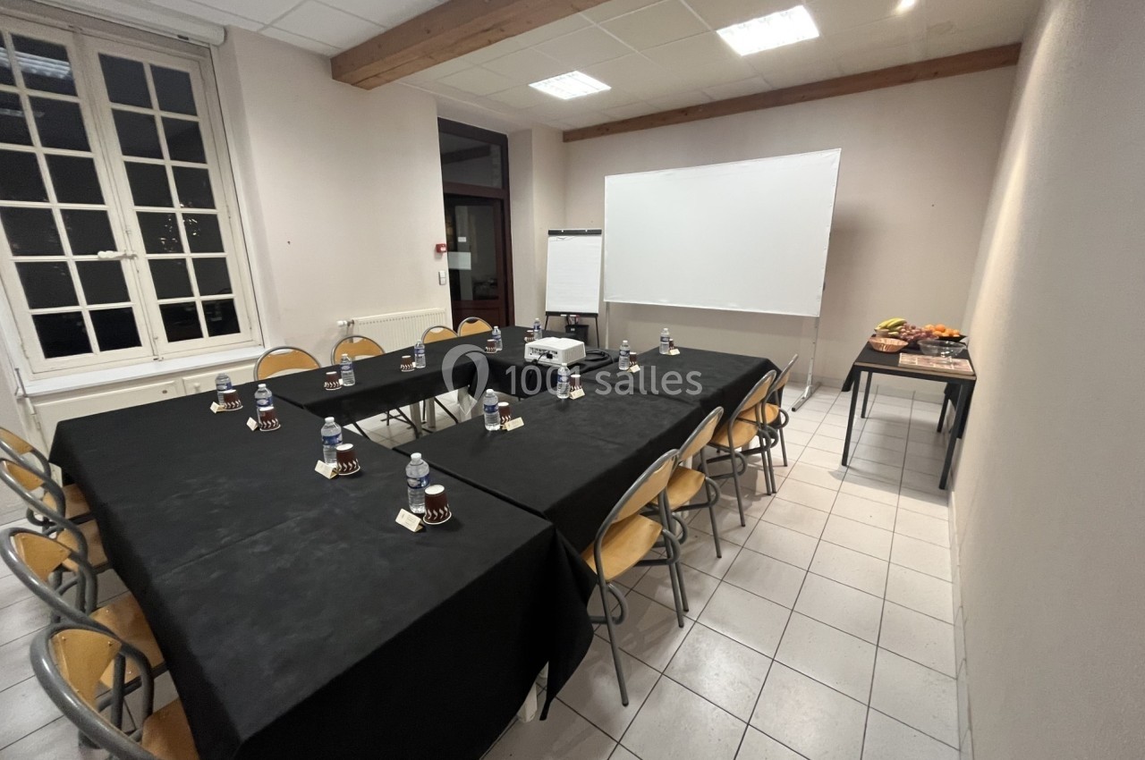 Salle de réunion avec table rectangulaire dressée, chaises, tableau blanc, vidéoprojecteur et collation sur une table.