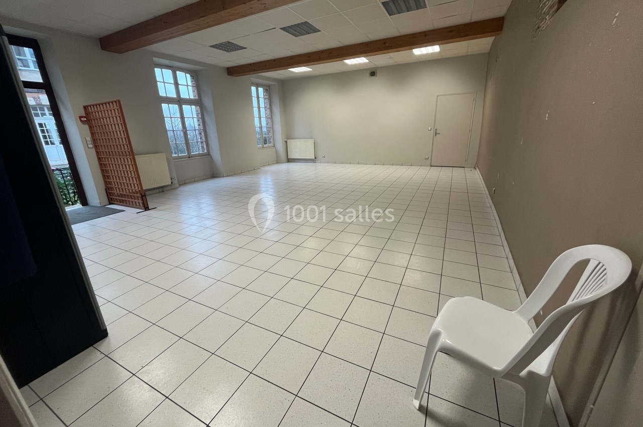 Salle vide avec carrelage blanc, murs beiges, plafond à poutres apparentes et une chaise en plastique blanc.