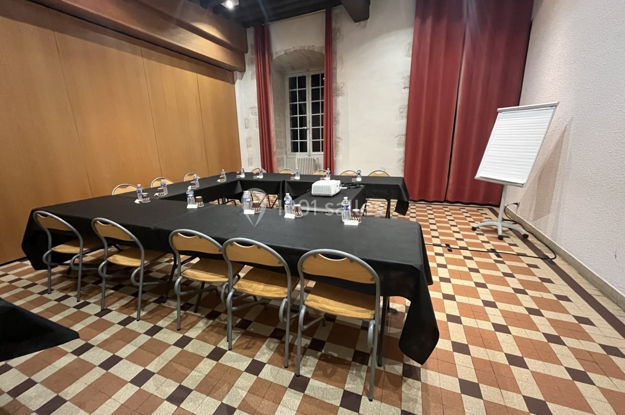 Salle de réunion avec tables noires disposées en U, chaises en bois, tableau de conférence et sol en carrelage.