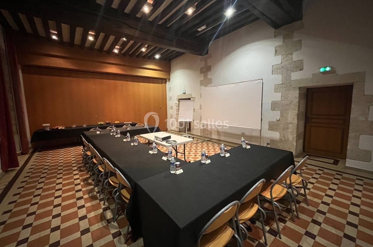 Salle de réunion avec tables disposées en U, nappes noires, chaises en bois, tableau blanc et éclairage tamisé.