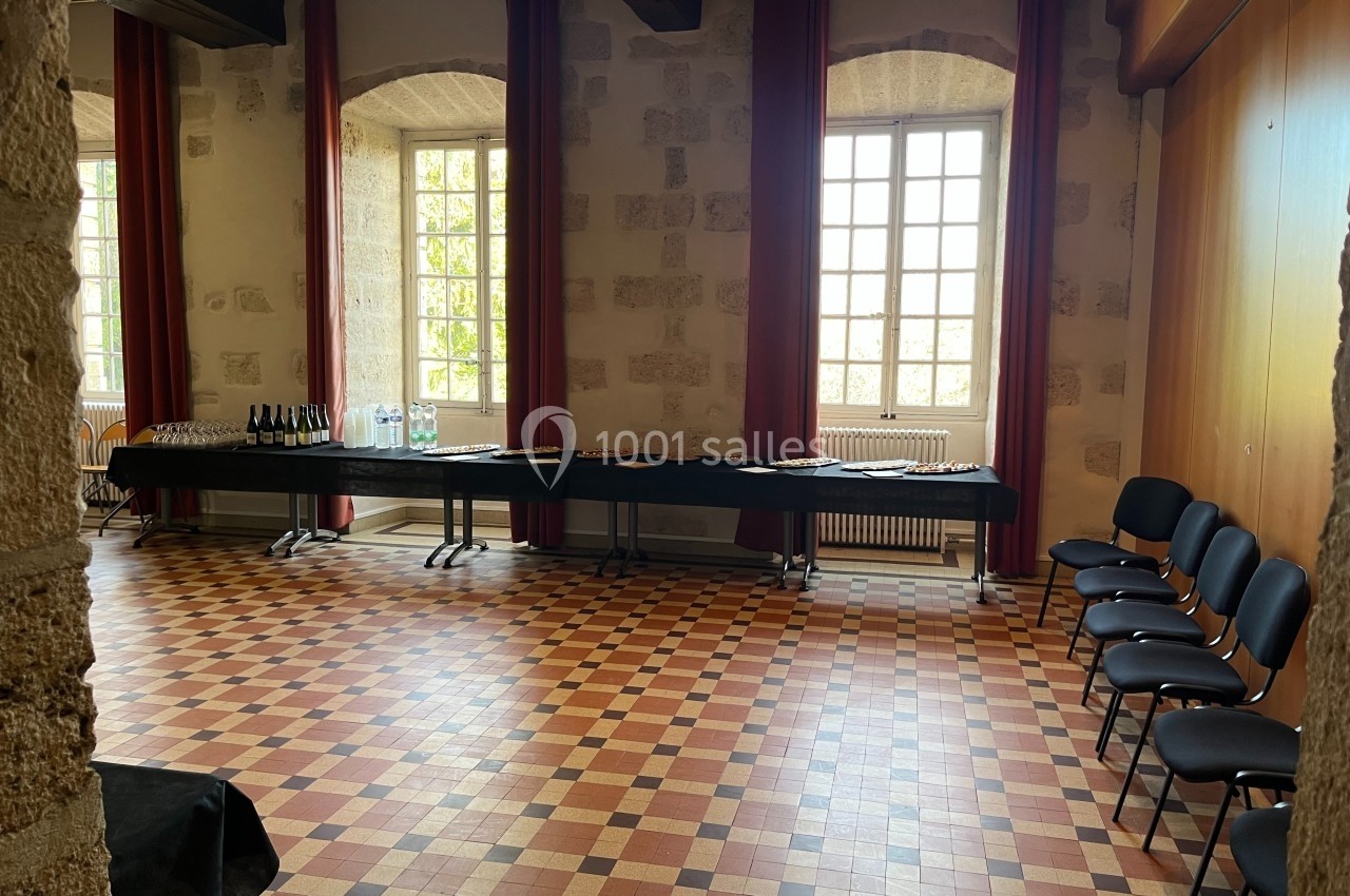 Salle lumineuse avec murs en pierre, sol en carreaux colorés, tables dressées avec boissons et chaises alignées.