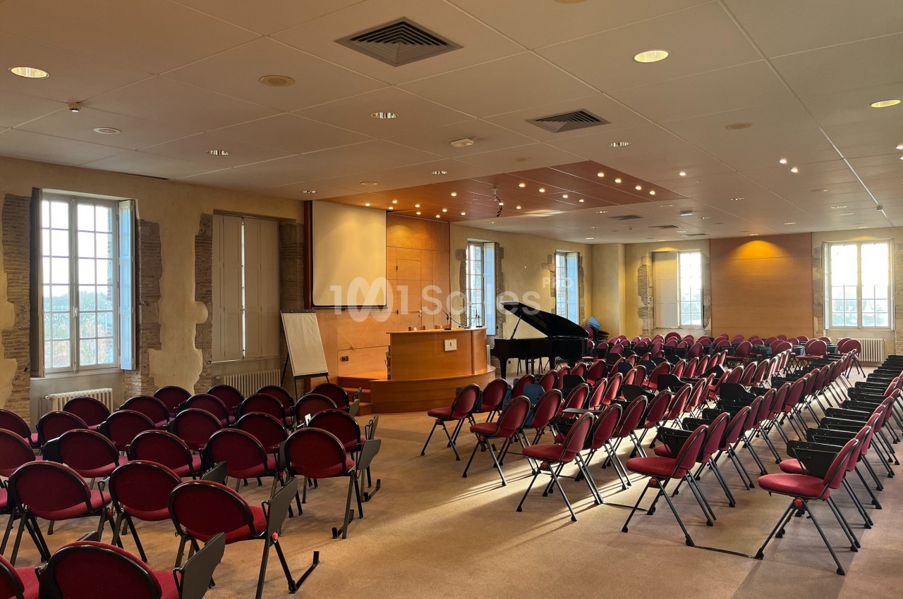 Salle de conférence lumineuse avec des rangées de chaises rouges, un piano et un pupitre en bois.