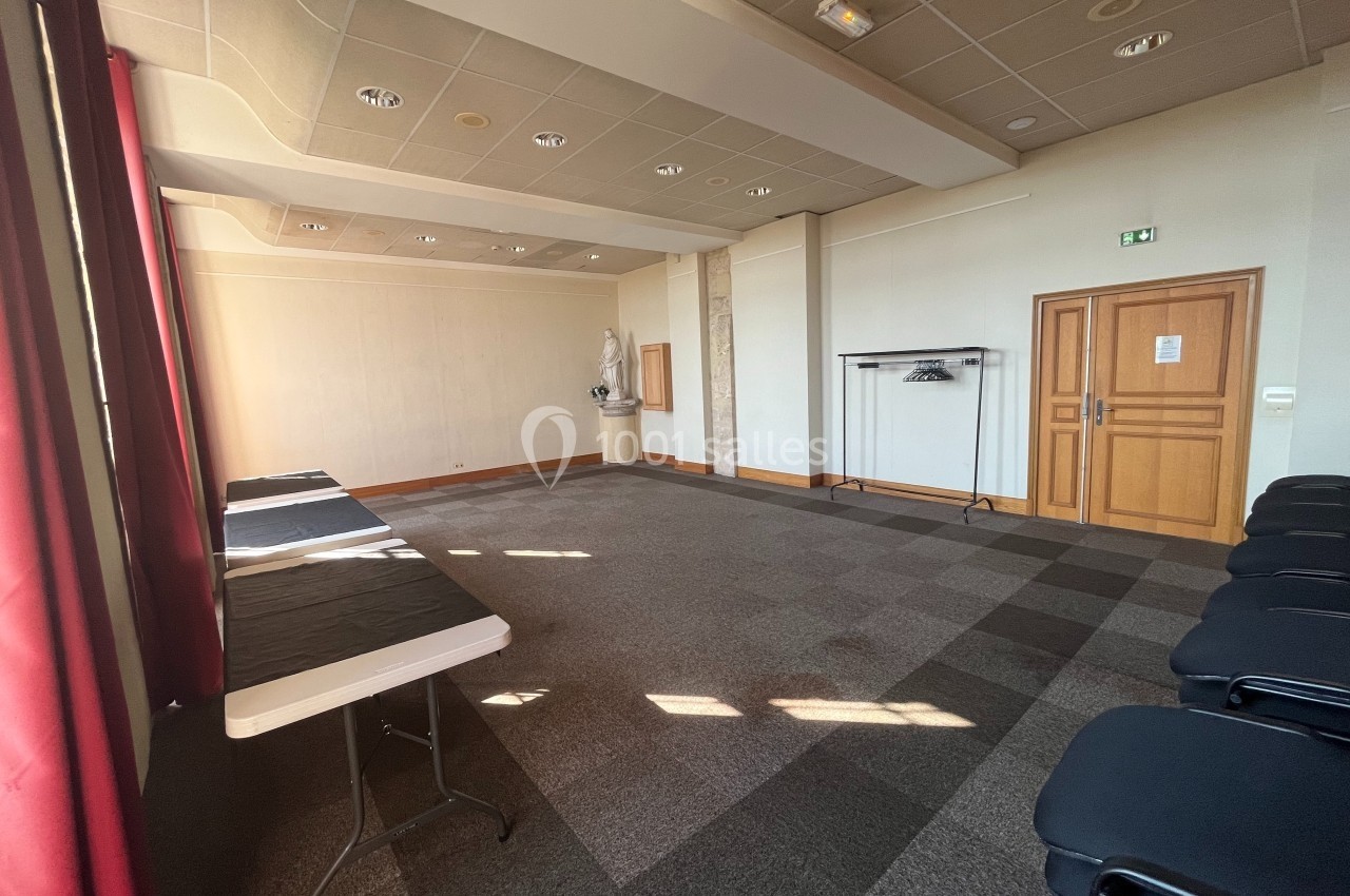 Salle lumineuse avec moquette grise, tables pliantes noires, chaises alignées et porte en bois au fond.