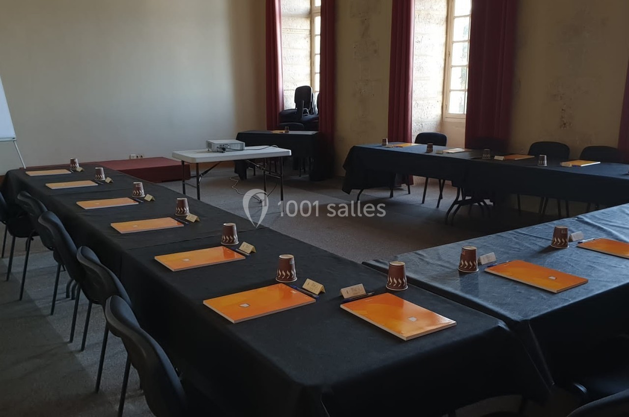 Salle de réunion avec tables disposées en U, équipées de dossiers, gobelets et un projecteur au fond de la pièce.