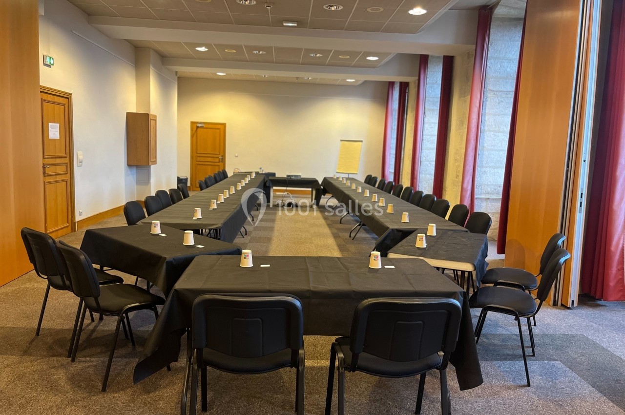 Salle de réunion avec tables disposées en U, nappes noires, chaises noires et gobelets alignés sur les tables.