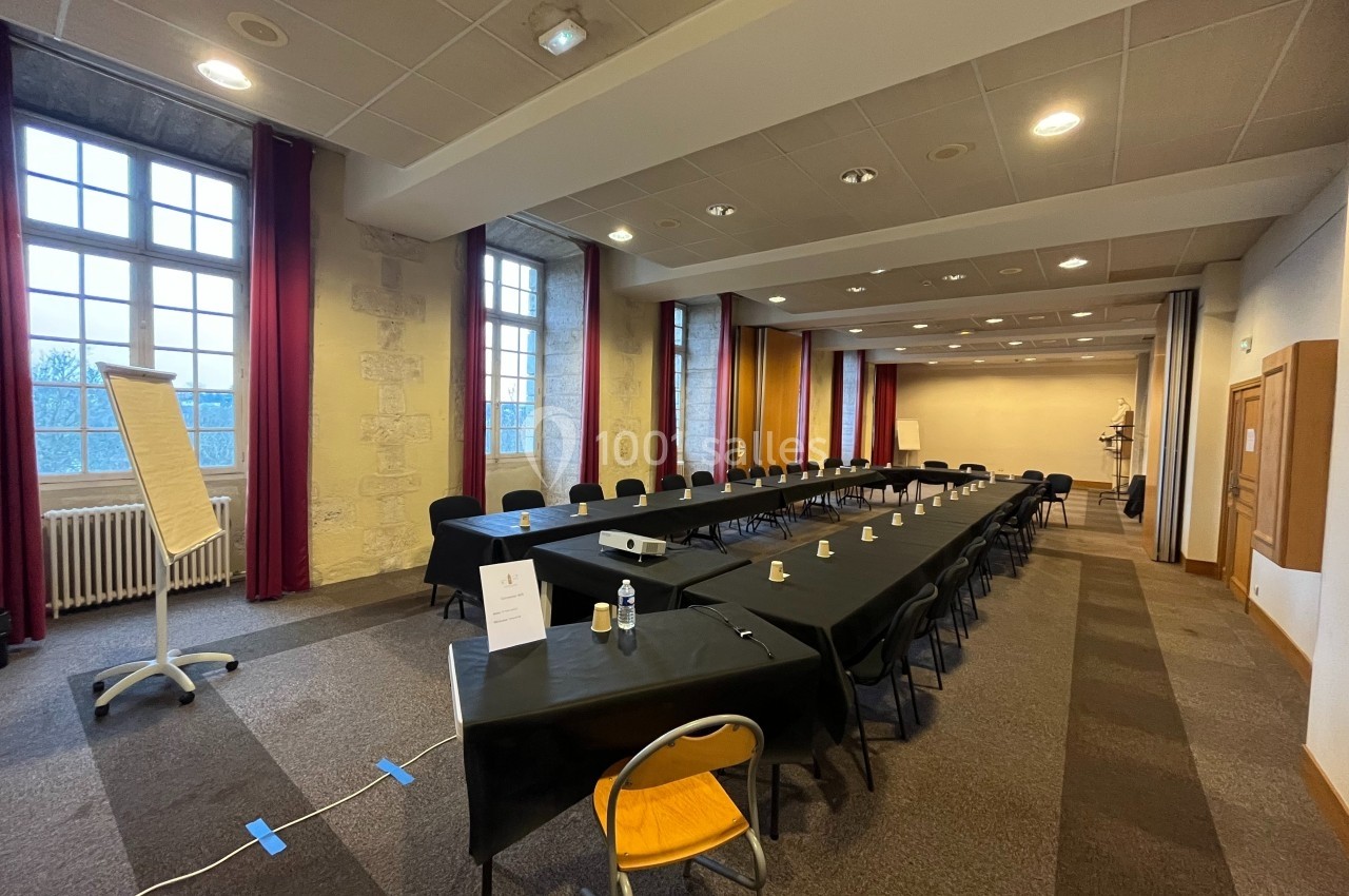 Salle de réunion avec tables disposées en U, chaises noires, tableau blanc et projecteur, éclairée par de grandes fenêtres.