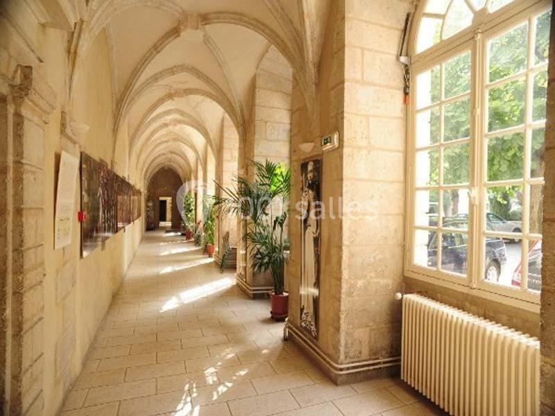 Un couloir voûté en pierre avec des plantes en pot, éclairé par la lumière naturelle entrant par de grandes fenêtres.