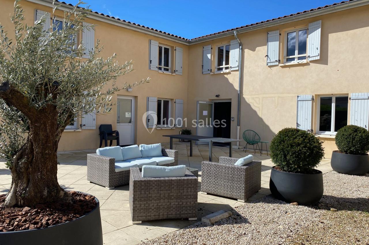 Cour intérieure avec mobilier d'extérieur en rotin, olivier et arbustes en pots, entourée d'un bâtiment beige à volets…