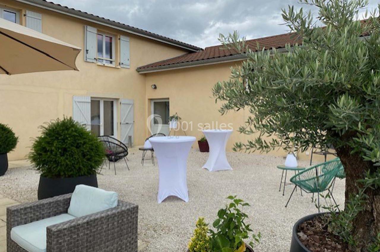 Cour extérieure avec mobilier de jardin, tables hautes drapées de blanc, plantes en pot et façade beige avec volets.