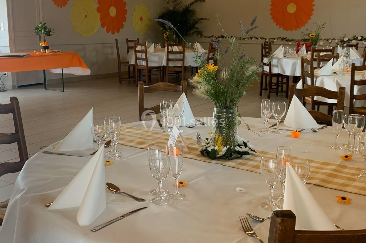 Salle décorée pour un événement, avec tables dressées, nappes blanches, serviettes pliées et décorations florales.