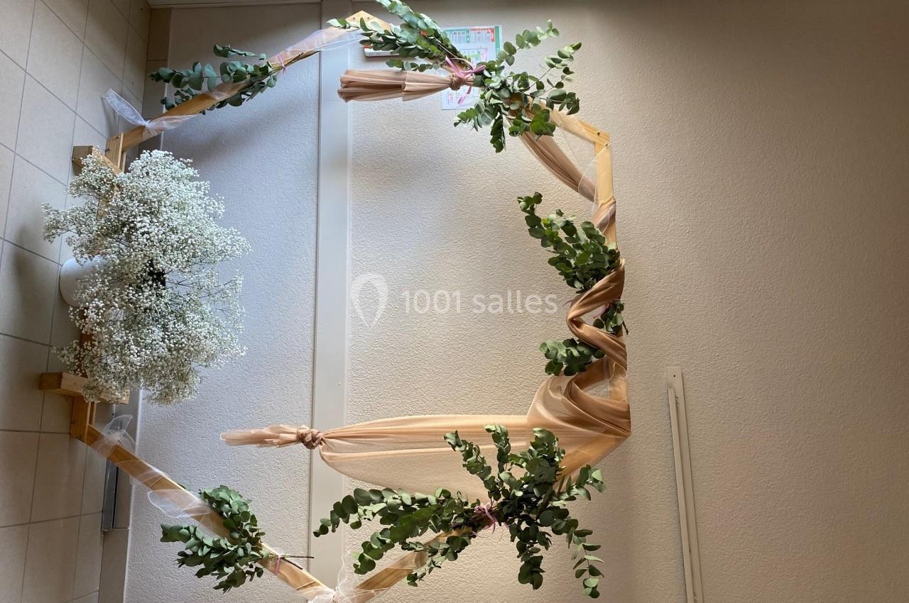 Arche décorative en bois ornée de feuillage, de gypsophile blanche et de voilages beige, placée dans un intérieur.