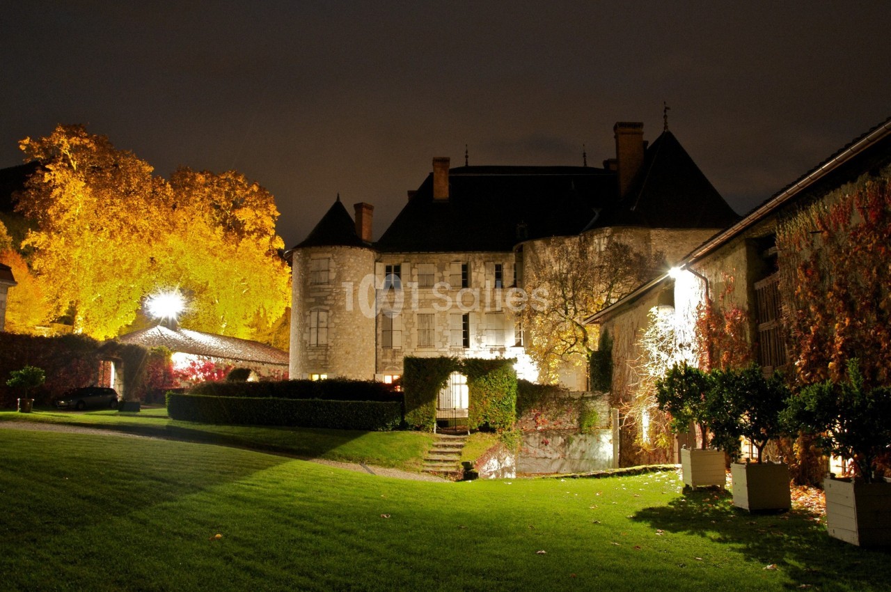 Château éclairé la nuit, entouré d'arbres aux feuilles automnales et d'une pelouse bien entretenue.