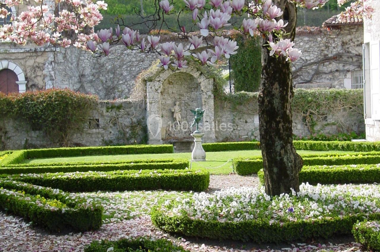Jardin avec parterres de buis, arbre en fleurs roses et statue au centre, entouré de murs en pierre.