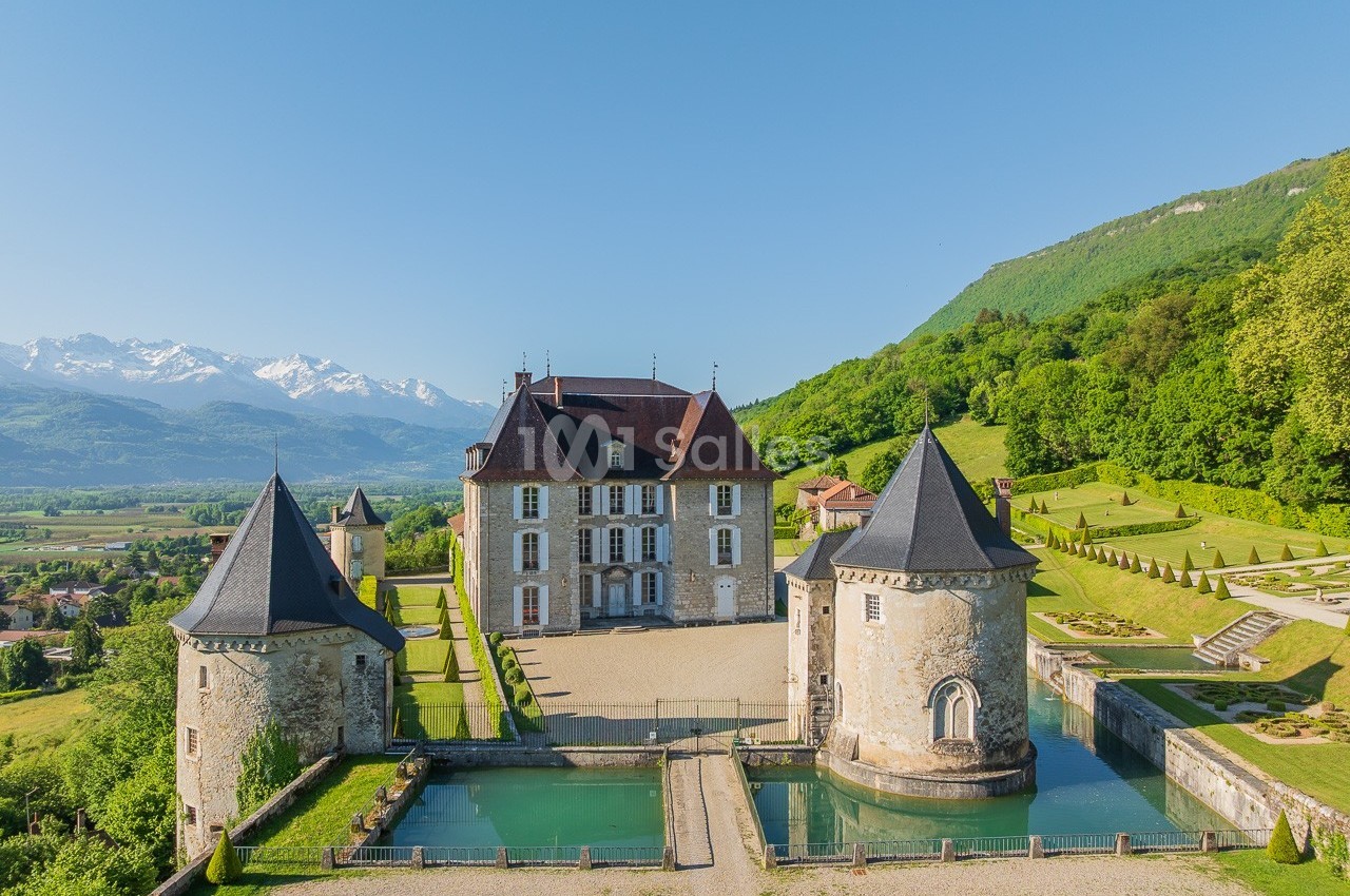 Vue aérienne d'un château entouré de douves, avec des montagnes enneigées et une forêt en arrière-plan.