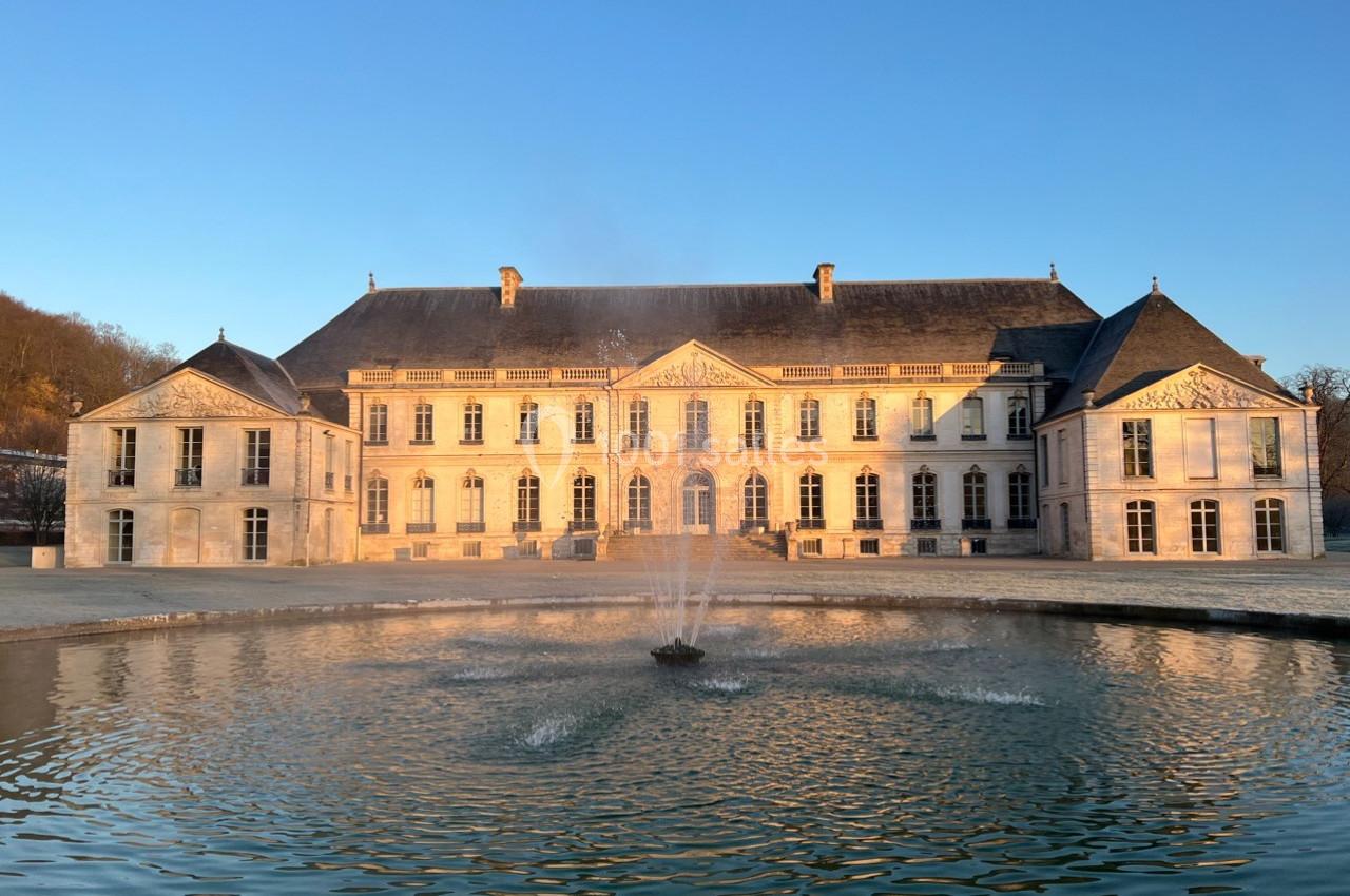 Façade d'un château éclairé par le soleil couchant, avec une fontaine au centre d'un bassin au premier plan.