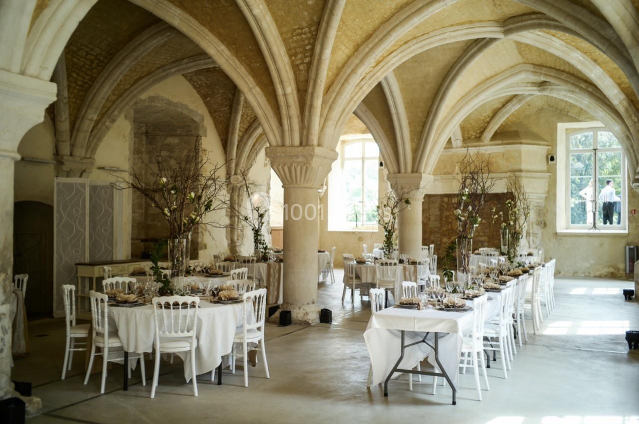 Salle voûtée avec tables élégamment dressées pour un événement, décorée de branches et baignée de lumière naturelle.