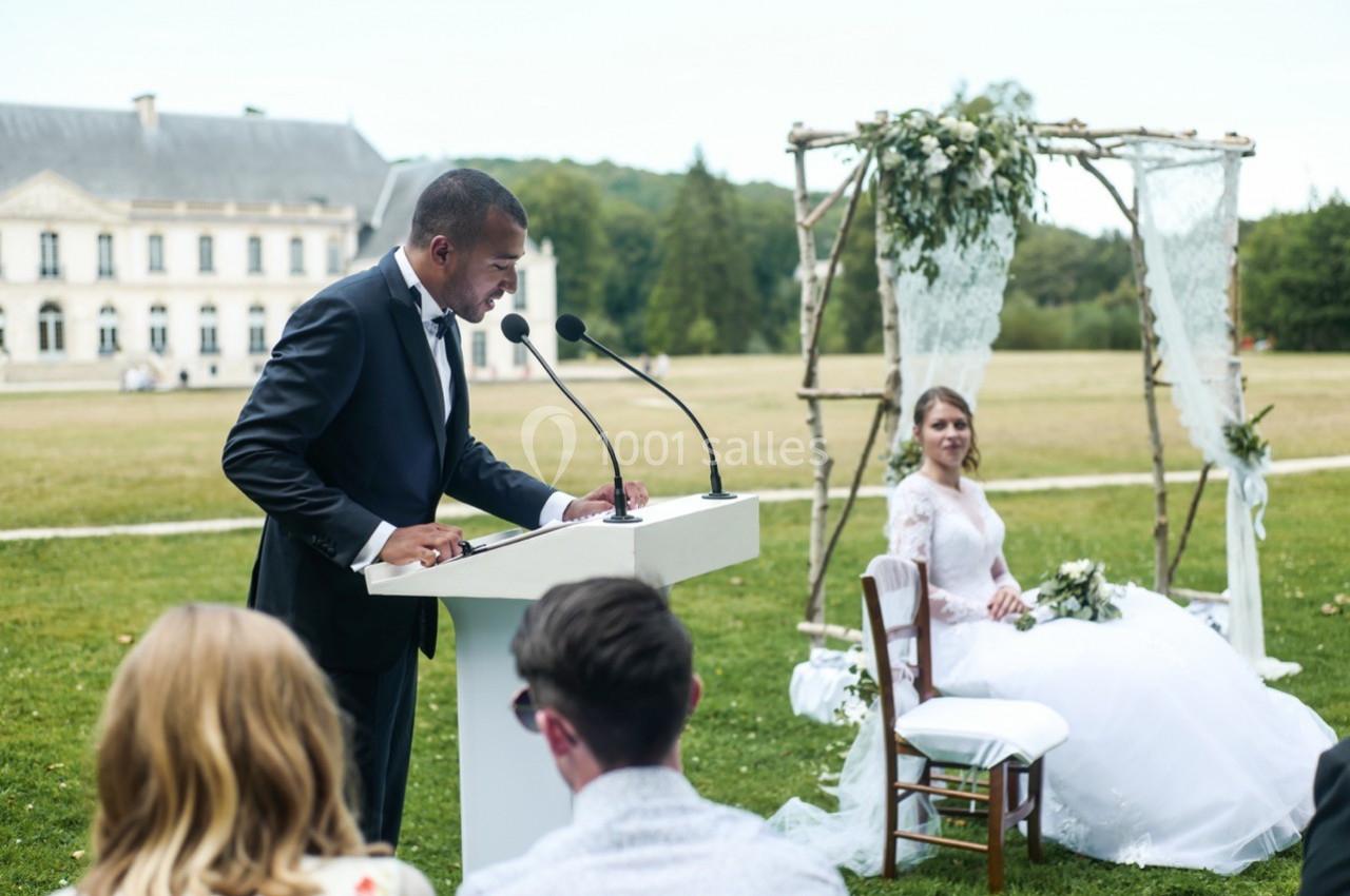 Un homme en costume prononce un discours devant un pupitre lors d'une cérémonie de mariage en extérieur.